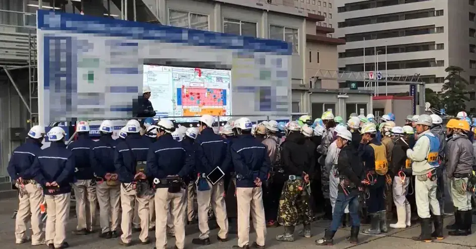 大型ビジョンの前で朝礼に集まった作業員
